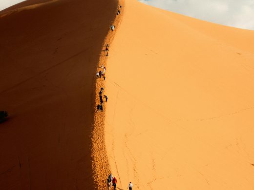 Silhouettes of people ascending a towering sand dune against a bright blue sky.