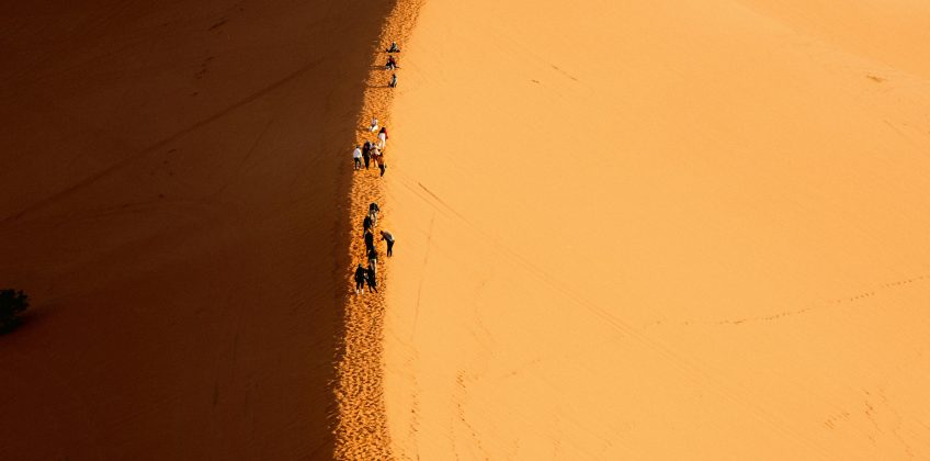 Silhouettes of people ascending a towering sand dune against a bright blue sky.